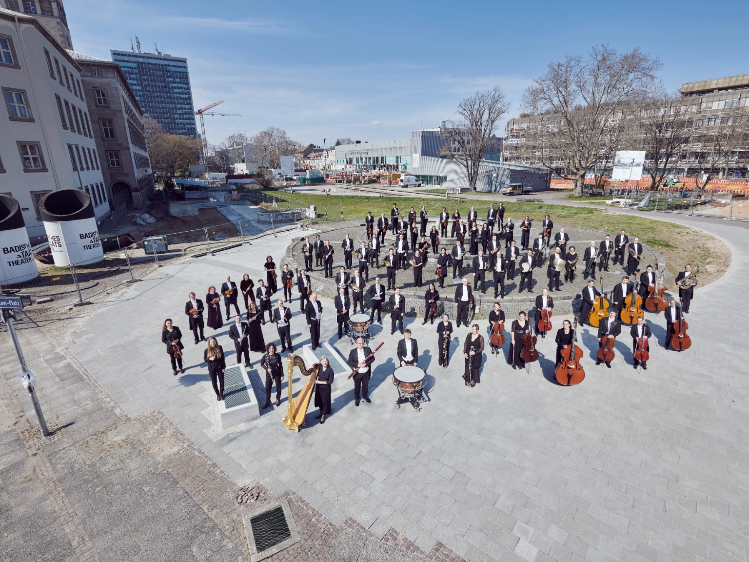 Ensemble Bild der Badischen Staatskapelle. Die Orchestermitglieder stehen gemeinsam dem Platz vor dem Badischen Staatstheater Karlsruhe. 