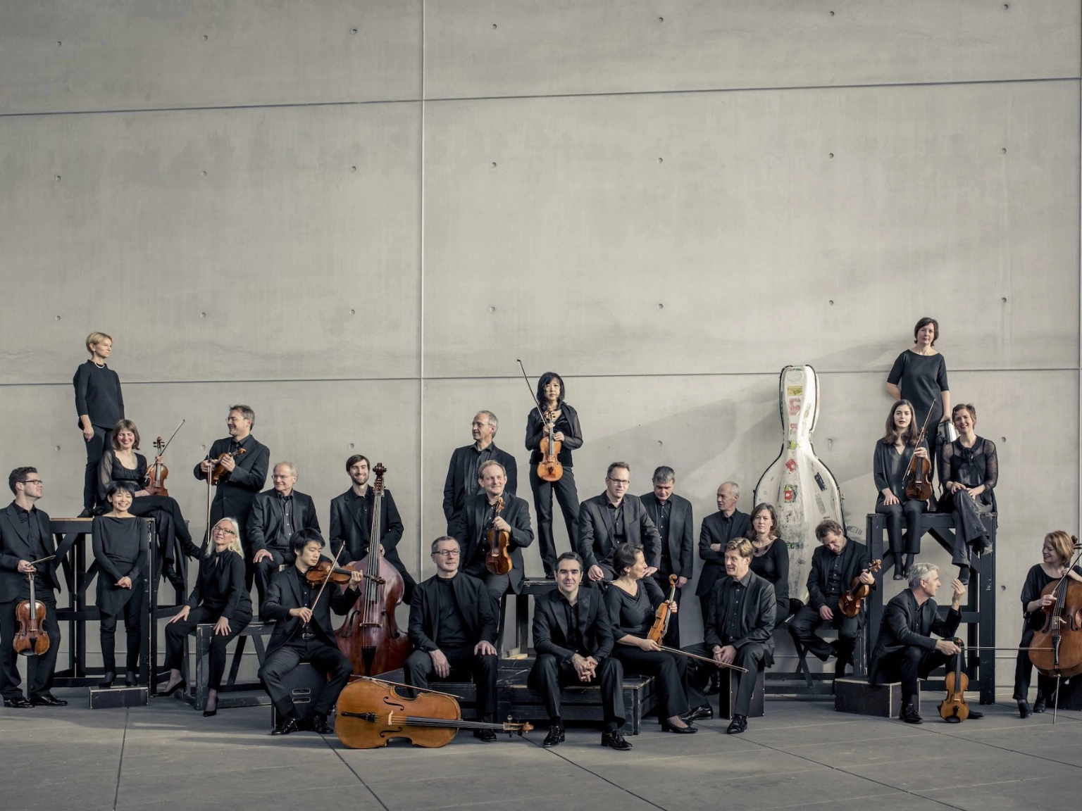 Das Münchener Kammerorchester vor einer grauen Betonwand. Sie posieren zusammen mit ihren Instrumenten. Manche schauen in die Kamera, andere unterhalten sich. 