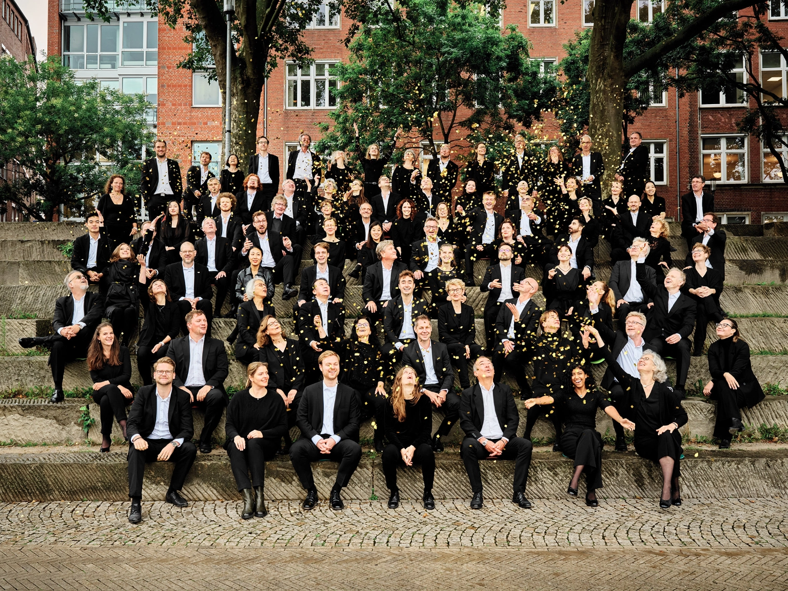 Orchesterbild Bremer Philharmoniker. <br/>Die Orchestermitglieder sitzen lachend auf Steinstufen unter Bäumen und werfen goldenes Konfetti in die Luft. 